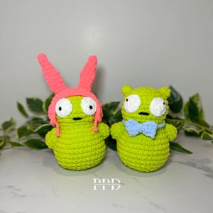Puede incluir: Dos juguetes de peluche verde hechos a crochet. Uno lleva un gorro de conejo rosa y el otro una pajarita azul.