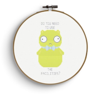 May include: Cross-stitch embroidery in a wooden hoop. The design features a lime green cartoon character with a blue bow tie and the text "DO YOU NEED TO USE THE FACILITIES?" on a white background.