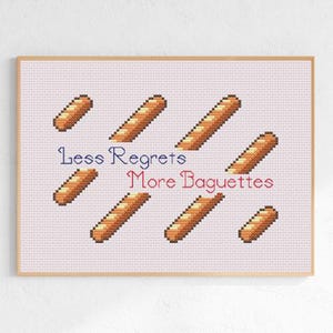 May include: Cross-stitch art print with pixelated baguettes in brown hues. The text "Less Regrets More Baguettes" is in blue and red. The artwork is framed in a light brown wooden frame.