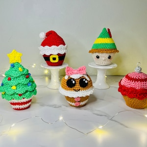 May include: Five crocheted Christmas-themed cupcakes. The cupcakes are decorated with Santa, a Christmas tree, a gingerbread person, an elf, and a pink and red ornament.