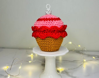 May include: A crocheted cupcake ornament with pink and red frosting and a brown base. The ornament is on a white cake stand with a string loop for hanging.