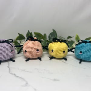 May include: Four crocheted plush figures in pastel colors: lavender, peach, yellow, and teal. Each has black yarn details for the eyes, mouth, arms, and a bow on top. The figures are arranged in a row against a white background with green leaves.