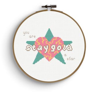May include: A cross-stitch embroidery hoop featuring a pink heart and teal star design with the words "you are stay gold a star". The embroidery is set against a white fabric background, framed by a wooden hoop.
