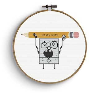 Op de afbeelding: Kruissteekborduurwerk in een houten hoepel met een stripfiguur die een potlood vasthoudt met de tekst "ME HOY MINOY". Het figuur heeft een vierkante vorm met grote ogen en een verbaasde uitdrukking. Het potlood is geel met een roze gum.