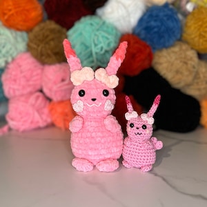 Puede incluir: Dos peluches de conejo de crochet rosa. El conejo más grande tiene un diseño de corazón rosa y blanco en la cabeza y un diseño de corazón rosa y blanco en las mejillas. El conejo más pequeño tiene un diseño de corazón rosa y blanco en la cabeza. Ambos conejos tienen ojos negros y una nariz rosa.