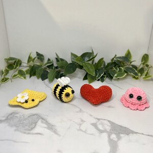 Puede incluir: Cuatro juguetes de peluche tejidos a crochet: una abeja amarilla, una flor amarilla, un corazón rojo y un pulpo rosa. Los juguetes están sobre una superficie de mármol blanco.