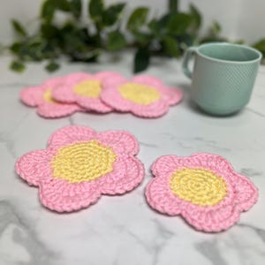 May include: Four crocheted pink flower coasters with yellow centers. The coasters are on a white marble surface.