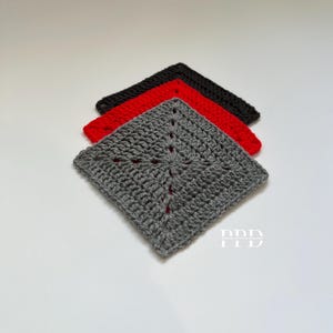 Pode incluir: Três quadrados de crochê em cinza, vermelho e preto. Os quadrados estão empilhados um em cima do outro, com o quadrado cinza no topo. Os quadrados têm um design geométrico simples.
