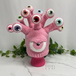May include: A handmade pink crochet creature with multiple eye stalks, each topped with a different colored eye. The main body has a large central eye. The creature is displayed on a pink base, with green foliage in the background.