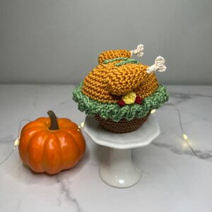 May include: A crocheted Thanksgiving turkey cupcake with a green and brown base, a yellow and red flower, and white legs. The cupcake is on a white cake stand next to a small orange pumpkin.