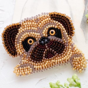 Può includere: Spilla a forma di carlino realizzata a mano con perline marroni, beige e nere. Il viso del carlino è dettagliato con occhi neri e naso scuro. La spilla è posta su uno sfondo bianco con un ritratto di cane ad acquerello.