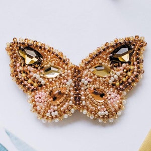 Peut inclure: Broche papillon perlée avec des perles dorées, marron et roses. Le papillon comporte de grands cristaux ambrés facettés. Les ailes sont ornées de petites perles dorées, roses et blanches.