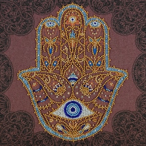 May include: A Hamsa hand wall art piece featuring intricate beadwork in gold, blue, and white. The design includes a central eye motif, floral patterns, and paisley shapes, all set against a textured dark mauve fabric background with a black border.