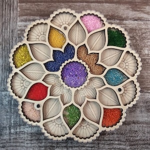 May include: A wooden bead organizer with 13 compartments shaped like flower petals. Each compartment is filled with colorful beads in various shades of pink, yellow, blue, green, red, and purple.