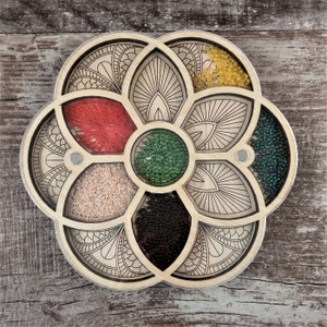 May include: A wooden bead organiser with seven compartments, each filled with different coloured beads. The organiser is shaped like a flower with a mandala design.