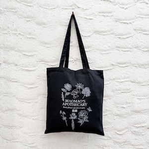May include: Black tote bag with white floral illustrations and text. The bag features the words "MAOMAO'S APOTHECARY" and the phrase "Every flower can cure or curse." The bag is hanging against a white brick wall.