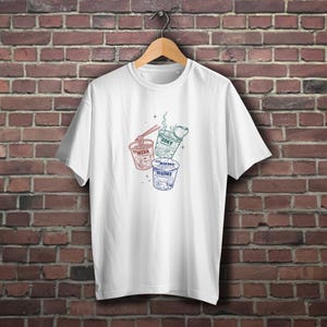 Peut inclure: T-shirt blanc avec un motif de trois bols de ramen, chacun portant un nom : "Mira", "Zoey" et "Rumi". Les bols sont rouges, verts et bleus, avec de la vapeur s'échappant du bol vert. Le t-shirt est suspendu à un cintre en bois.