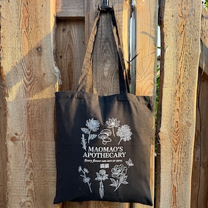 May include: Black tote bag with white floral illustrations and text. The text reads "MAOMAO'S APOTHECARY" and "Every flower can cure or curse" with Chinese characters. The bag is hanging on a wooden fence.