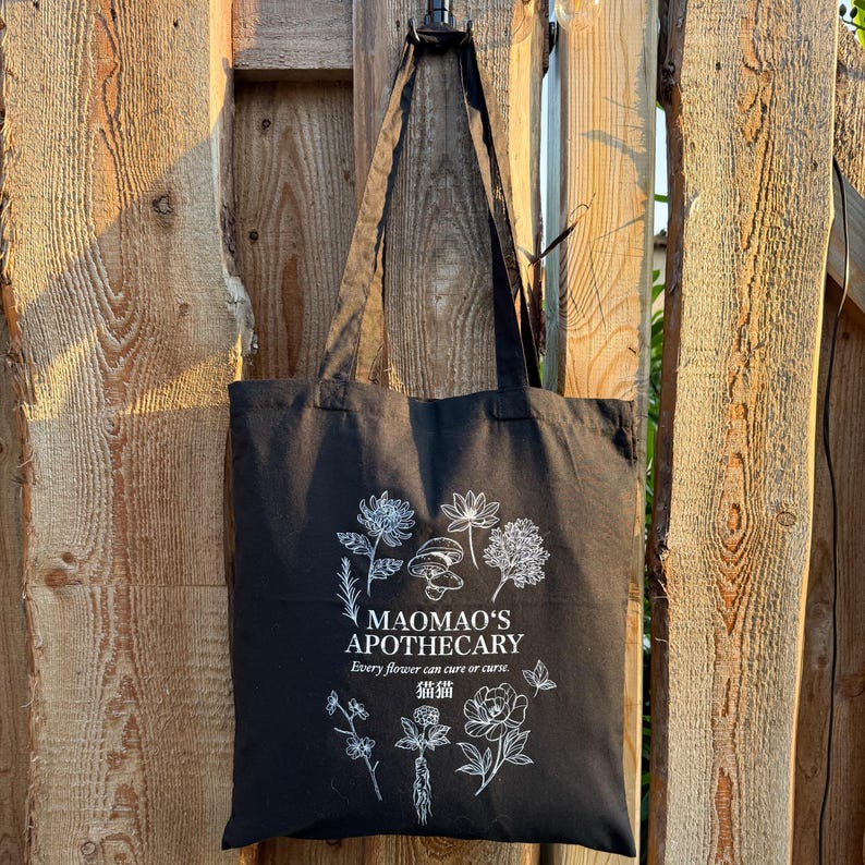 May include: Black tote bag with white floral illustrations and text. The text reads "MAOMAO'S APOTHECARY" and "Every flower can cure or curse" with Chinese characters. The bag is hanging on a wooden fence.