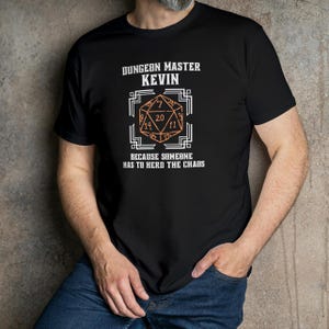 Könnte beinhalten: Schwarzes T-Shirt mit dem Text "DUNGEON MASTER KEVIN" über einem geometrischen Design mit den Zahlen 7, 11, 14 und 20. Darunter steht der Text "BECAUSE SOMEONE HAS TO HERD THE CHAOS".