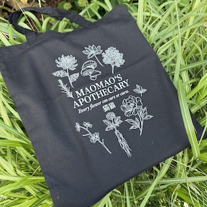 May include: Black tote bag with white botanical illustrations and text. The bag features the words "MAOMAO'S APOTHECARY" and the phrase "Every flower can cure or curse." The design includes drawings of flowers, mushrooms, and other plants.
