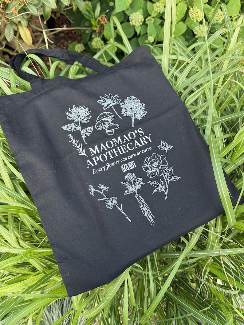 May include: Black tote bag with white botanical illustrations and text. The bag features the words "MAOMAO'S APOTHECARY" and the phrase "Every flower can cure or curse." The design includes drawings of flowers, mushrooms, and other plants.