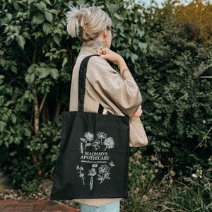 May include: Black tote bag with white floral and mushroom illustrations. The text "MAOMAO'S APOTHECARY" is printed above the floral design, with additional text below. The bag has black handles and is carried over the shoulder.