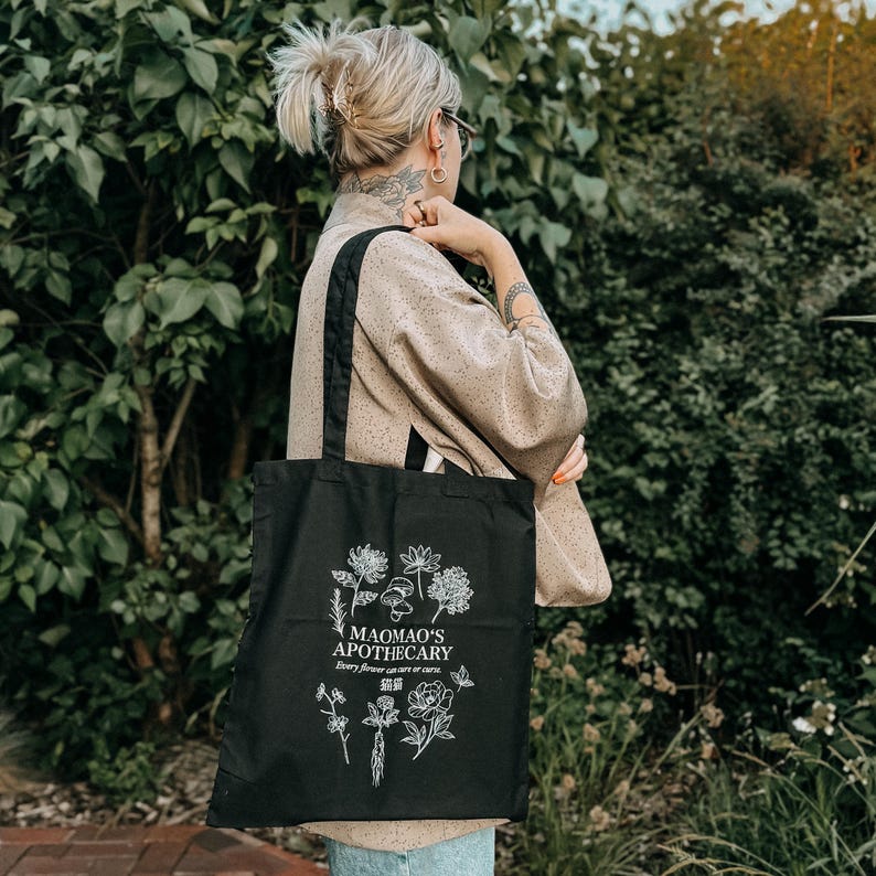 May include: Black tote bag with white floral and mushroom illustrations. The text "MAOMAO'S APOTHECARY" is printed above the floral design, with additional text below. The bag has black handles and is carried over the shoulder.