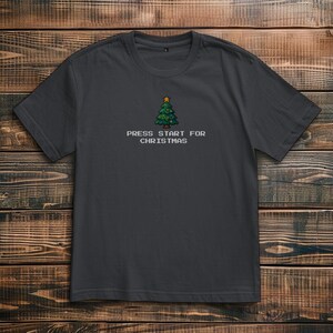Könnte beinhalten: Dunkelgraues T-Shirt mit einem pixeligen Weihnachtsbaum-Motiv und dem Text "PRESS START FOR CHRISTMAS". Der Baum ist grün mit einem gelben Stern. Das Shirt ist aus weichem Material.