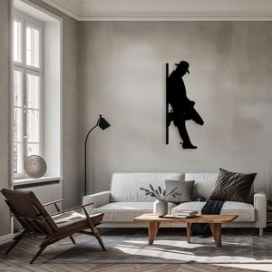 Pu&ograve; includere: Un'opera d'arte da parete in metallo nero a forma di silhouette di una persona appoggiata a un muro, con un cappello e un lungo cappotto. L'opera &egrave; esposta in un soggiorno con un divano bianco, un tavolino da caff&egrave; in legno e una lampada da terra.