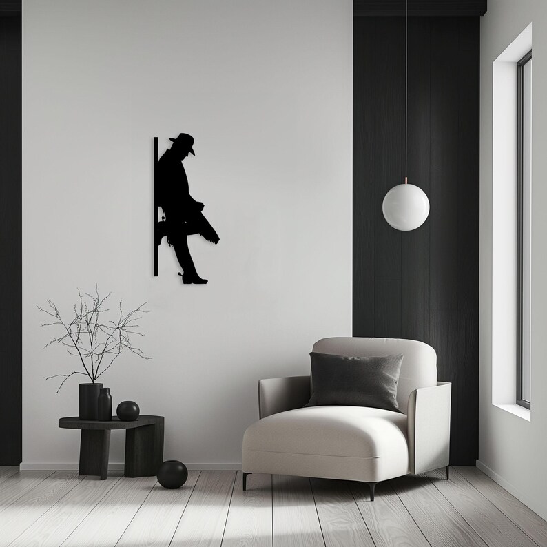 Pu&ograve; includere: Silhouette nera di una persona seduta su uno spigolo, con un cappello e che tiene una valigetta. La silhouette &egrave; montata su una parete bianca, con una poltrona bianca e un tavolino con un vaso e una pianta in primo piano.