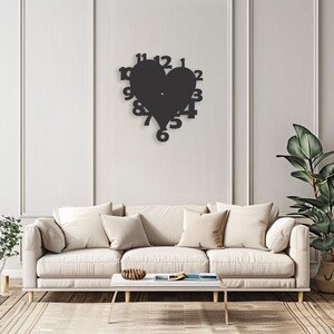 May include: Black heart-shaped wall clock with numbers around the edge. The clock is mounted on a white wall above a white couch with pillows.