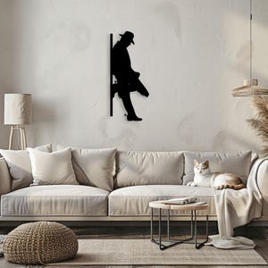 Pu&ograve; includere: Arte da parete in metallo nero a forma di silhouette di una persona con un cappello che si appoggia a un muro. La silhouette &egrave; montata su una parete bianca in un soggiorno.