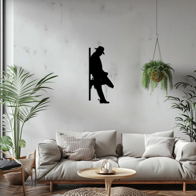 Pu&ograve; includere: Arte da parete in metallo nero a silhouette che rappresenta un cowboy seduto su un palo di recinzione. L'opera &egrave; montata su una parete bianca in un ambiente da soggiorno.