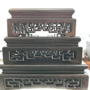 May include: Three dark brown wooden display stands stacked on top of each other. Each stand has a rectangular shape with a carved, openwork design on the sides. The stands are likely for displaying small objects.