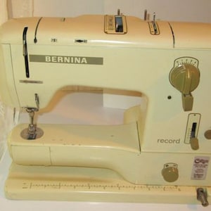 May include: A vintage white Bernina sewing machine with a gold-coloured dial and a record button. The machine has a built-in needle threader and a foot pedal.