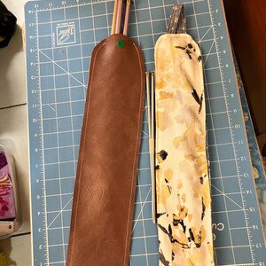 May include: Two pencil cases are displayed on a blue cutting mat. One is brown leather with a green button closure, and the other is a floral fabric with yellow and black accents. Pencils and pens are visible inside the cases.