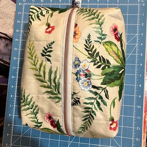 Op de afbeelding: Een rechthoekig tasje met rits van crèmekleurige stof met een botanische print van groene varens en kleine rode, gele en blauwe bloemen. Het tasje heeft een koperkleurige ritssluiting in het midden en ligt op een blauwe snijmat.
