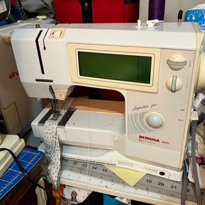 May include: A white and beige Bernina 1630 Inspiration Plus sewing machine featuring a digital display screen, two circular control knobs, a central button, and a needle currently stitching a piece of white fabric with a small blue floral pattern.