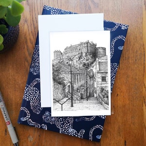 Può includere: Un disegno a penna e inchiostro in bianco e nero di un castello su una collina, con una scena di strada in primo piano. Il disegno è su una carta bianca, appoggiata su un taccuino blu navy con un motivo bianco. Sono visibili anche una penna nera e una piccola pianta verde.