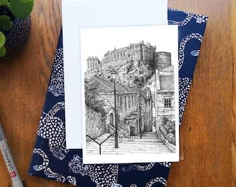 Puede incluir: Un dibujo a pluma y tinta en blanco y negro de un castillo en una colina, con una escena de calle en primer plano. El dibujo está sobre una tarjeta blanca, apoyada en un cuaderno azul marino con un patrón blanco. También se ve un bolígrafo negro y una pequeña planta verde.