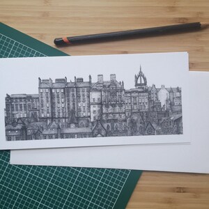 Puede incluir: Dibujo a tinta en blanco y negro de un paisaje urbano, con edificios y tejados detallados. La obra de arte est&aacute; sobre papel blanco, con un l&aacute;piz cerca. La imagen est&aacute; sobre una esterilla de corte verde y una superficie de madera.