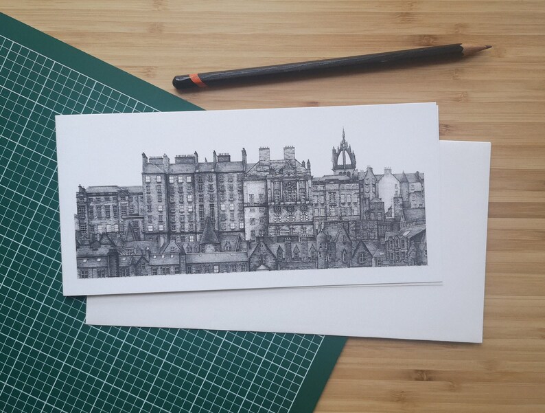 Puede incluir: Dibujo a tinta en blanco y negro de un paisaje urbano, con edificios y tejados detallados. La obra de arte est&aacute; sobre papel blanco, con un l&aacute;piz cerca. La imagen est&aacute; sobre una esterilla de corte verde y una superficie de madera.