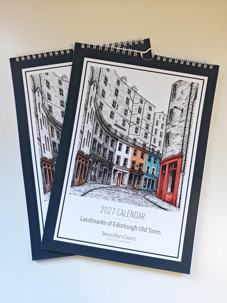 May include: Two spiral-bound 2027 calendars titled Landmarks of Edinburgh Old Town by Jennifer Court. The cover features a black and white ink illustration of city buildings with selective orange and blue color accents on the storefronts.