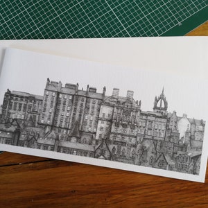 Puede incluir: Un dibujo a pluma en blanco y negro de un paisaje urbano, con edificios y tejados detallados. La obra de arte est&aacute; sobre una tarjeta blanca, con un sobre a juego. La imagen est&aacute; sobre una esterilla de corte verde y una superficie de madera.