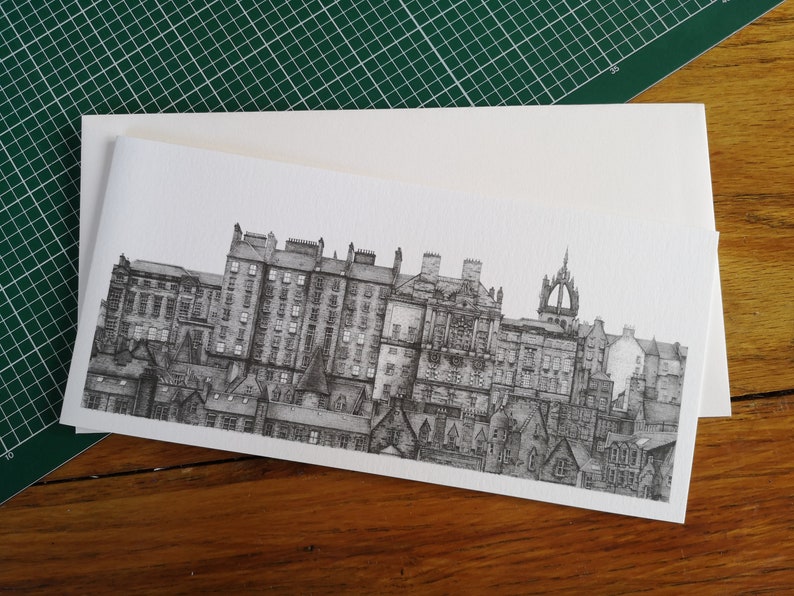 Puede incluir: Un dibujo a pluma en blanco y negro de un paisaje urbano, con edificios y tejados detallados. La obra de arte est&aacute; sobre una tarjeta blanca, con un sobre a juego. La imagen est&aacute; sobre una esterilla de corte verde y una superficie de madera.