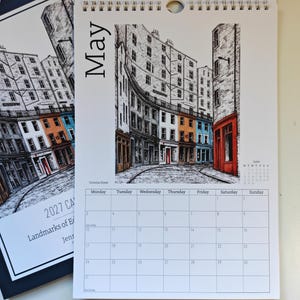 May include: A wall calendar page for May 2027 featuring a black and white ink illustration of Victoria Street in Edinburgh with colorful building facades in orange, blue, and red. The calendar grid below shows days of the week and dates.