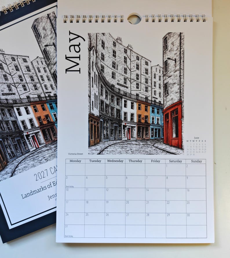 May include: A wall calendar page for May 2027 featuring a black and white ink illustration of Victoria Street in Edinburgh with colorful building facades in orange, blue, and red. The calendar grid below shows days of the week and dates.