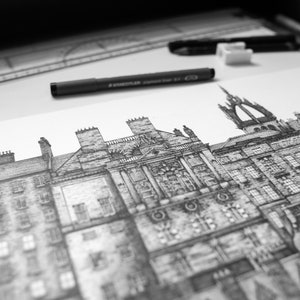 Puede incluir: Dibujo arquitect&oacute;nico en blanco y negro de un paisaje urbano detallado, con edificios de fachadas intrincadas, chimeneas y una torre prominente. Un bol&iacute;grafo negro, una goma de borrar y una regla est&aacute;n sobre la superficie blanca.