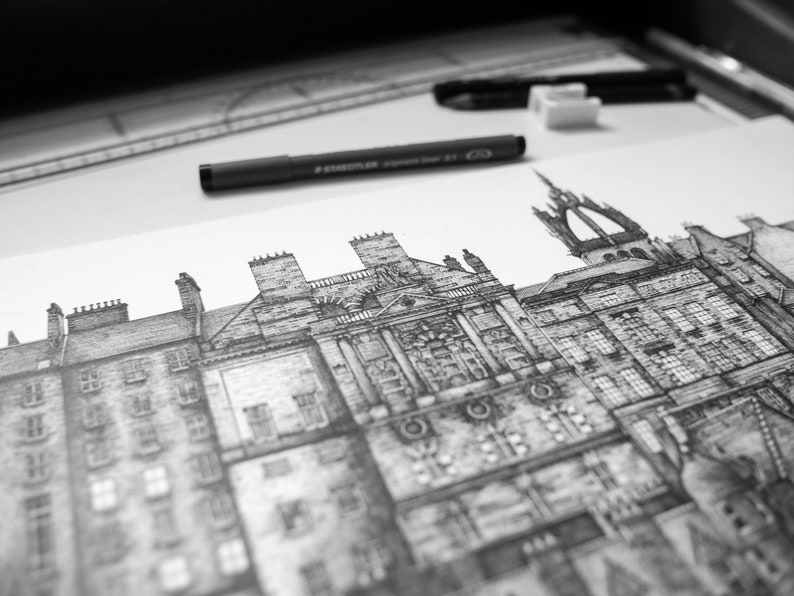 Puede incluir: Dibujo arquitect&oacute;nico en blanco y negro de un paisaje urbano detallado, con edificios de fachadas intrincadas, chimeneas y una torre prominente. Un bol&iacute;grafo negro, una goma de borrar y una regla est&aacute;n sobre la superficie blanca.